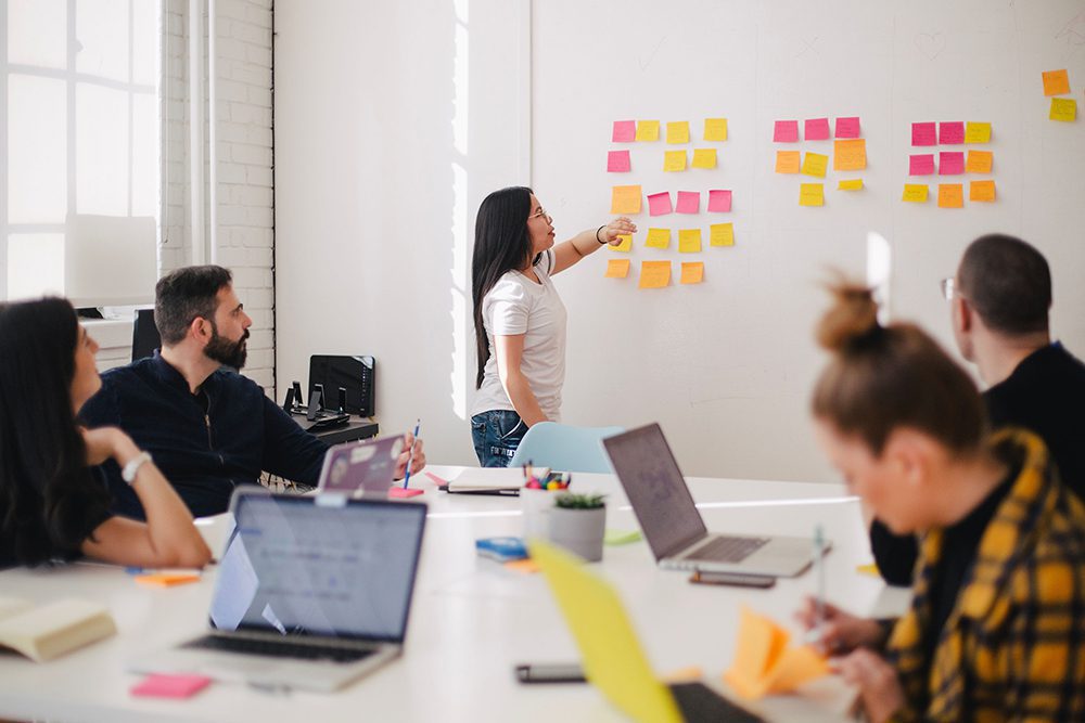 People sitting around a table, listening while woman gives presentation on wall in front of them