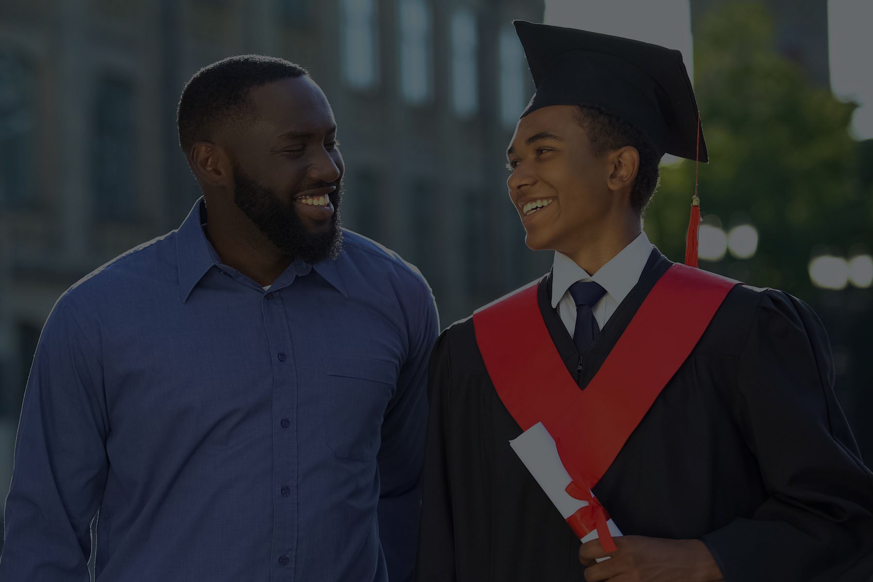 A father and son looking at each other smiling. The son has a graduation uniform and diploma in his hand