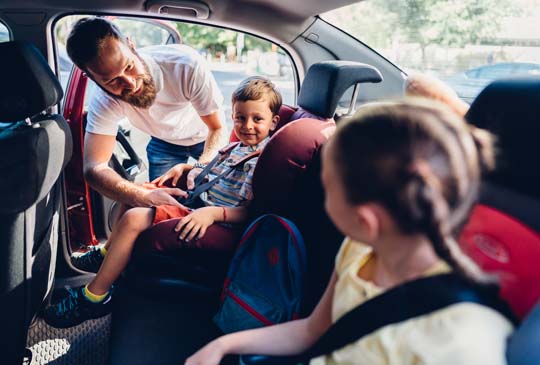 Dad buckling a little boy into his car seat. The little boy is smiling a the little girl next to him who is also in a car seat