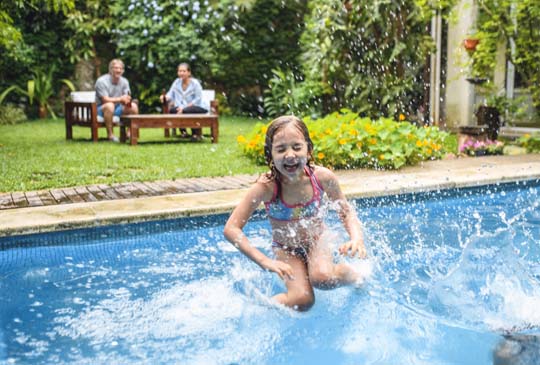 A young girl laughing and splashing into the backyard swimming pool with adults sitting in the background