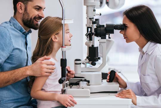 A women giving a little girl an eye exam through a phoroptor. The little girl is supported by a male figure behind her smiling and holding her arm