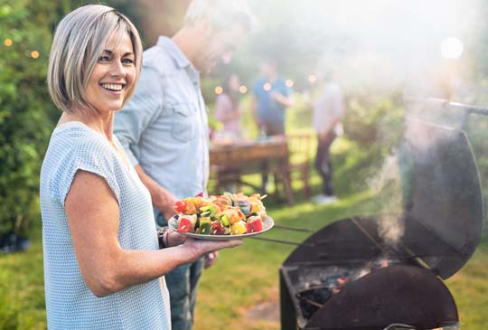 A women smiling and holding a platter of food while a man behind her is cooking on the grill