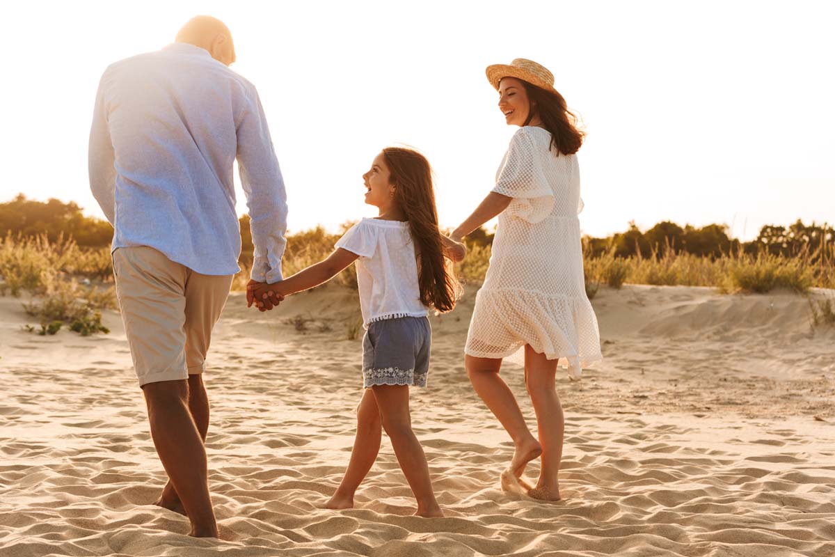 A mom, dad, and little girl holding hands and walking along the sand smiling at each other