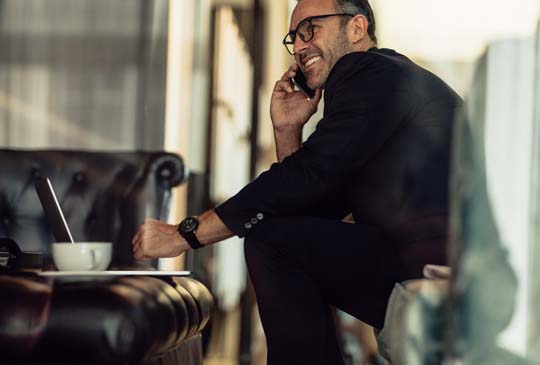 A man in a suit smiling on the phone and looking into the distance while sitting and enjoying coffee with his computer in front of him