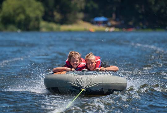 Two kids tubing in the water with smiles on their face and a rope pulling them
