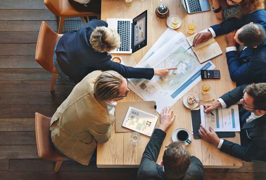 People sitting at a table with their computers and paper. One person is pointing to something on a piece of paper