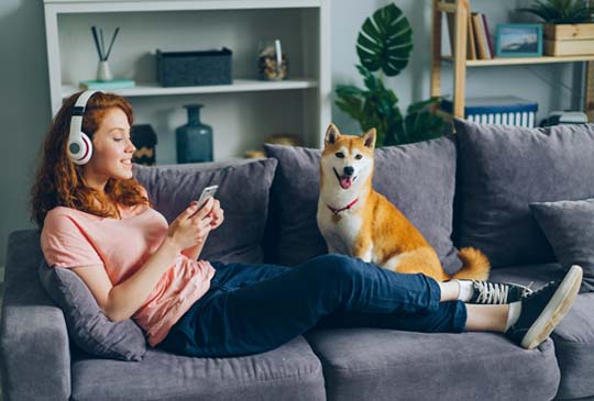 Women with headphones on and phone in hand sitting down on a couch with a dog sitting beside her