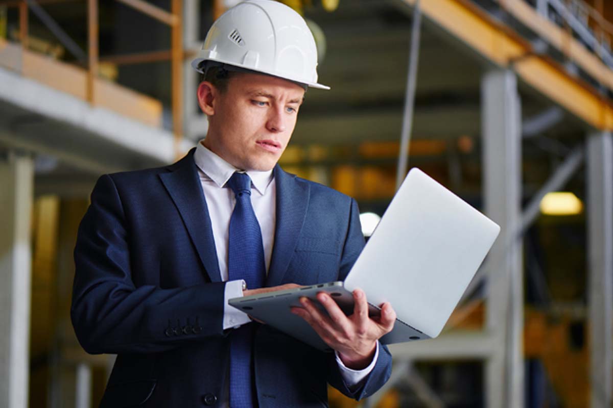 A man in a suit with a construction hat on standing and typing on a laptop