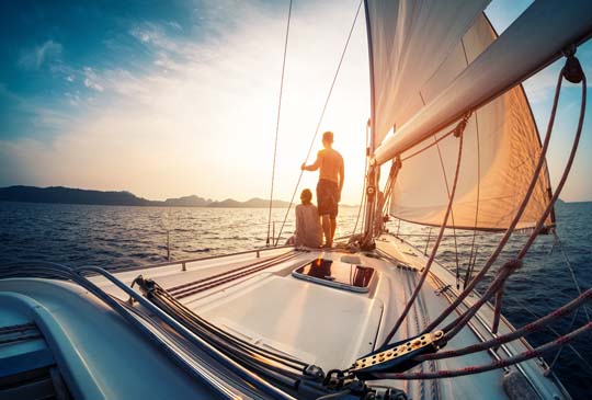 One person sitting on a sailboat in the water while the other is standing as he holds onto a rope looking into the distance