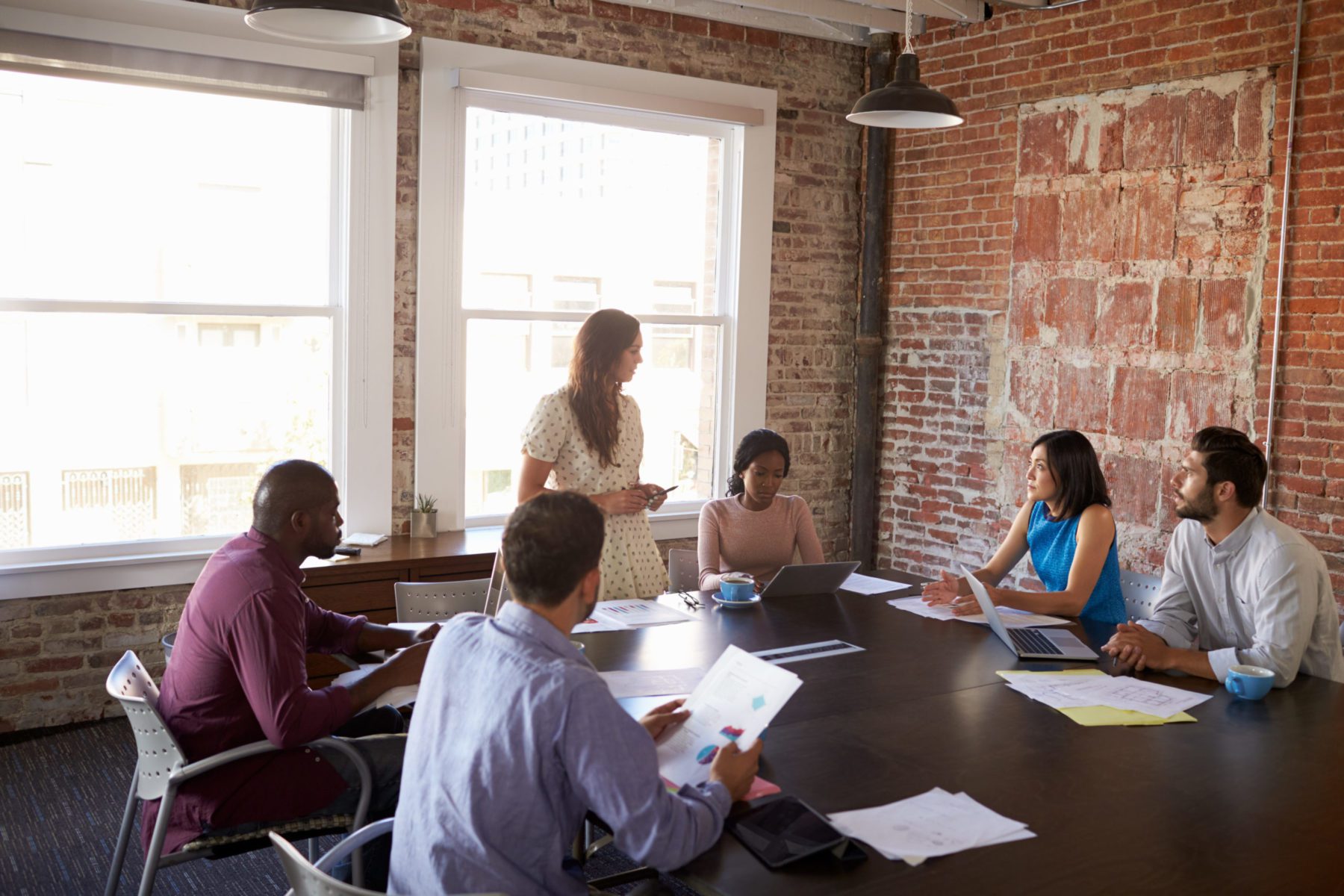 Six people at a meeting gathered around a table with papers and laptops