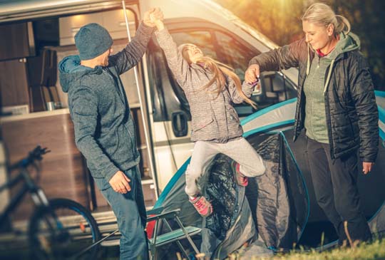 Mother and father swinging their daughter with their arms outside of the RV with a tent and bicycle in the background