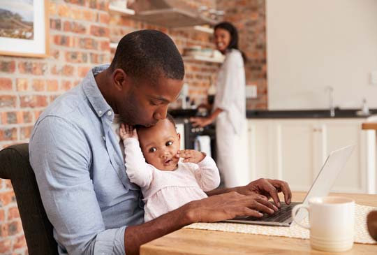 A little girl sitting on her dads lap as he types on his computer at the table. The mom is featured in the background smiling at them from the kitchen