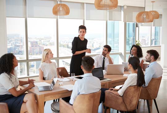 A lady standing and talking to a group of business people who are sitting and looking up at her around a table filled with laptops and papers