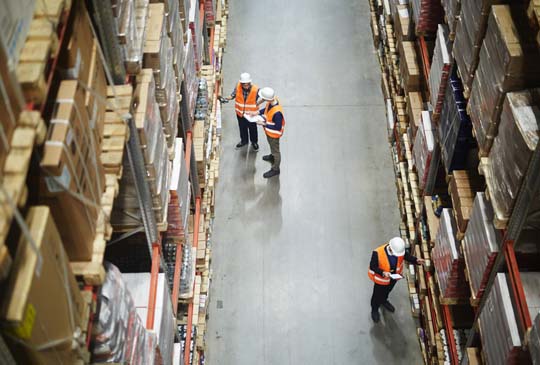 An overview of workers at a warehouse. There are three workers looking at the shelves and checking their clipboards