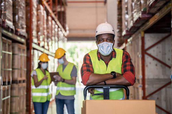 Man in a warehouse with his uniform and mask on looking at the camera. In the background there are two workers with masks on talking.